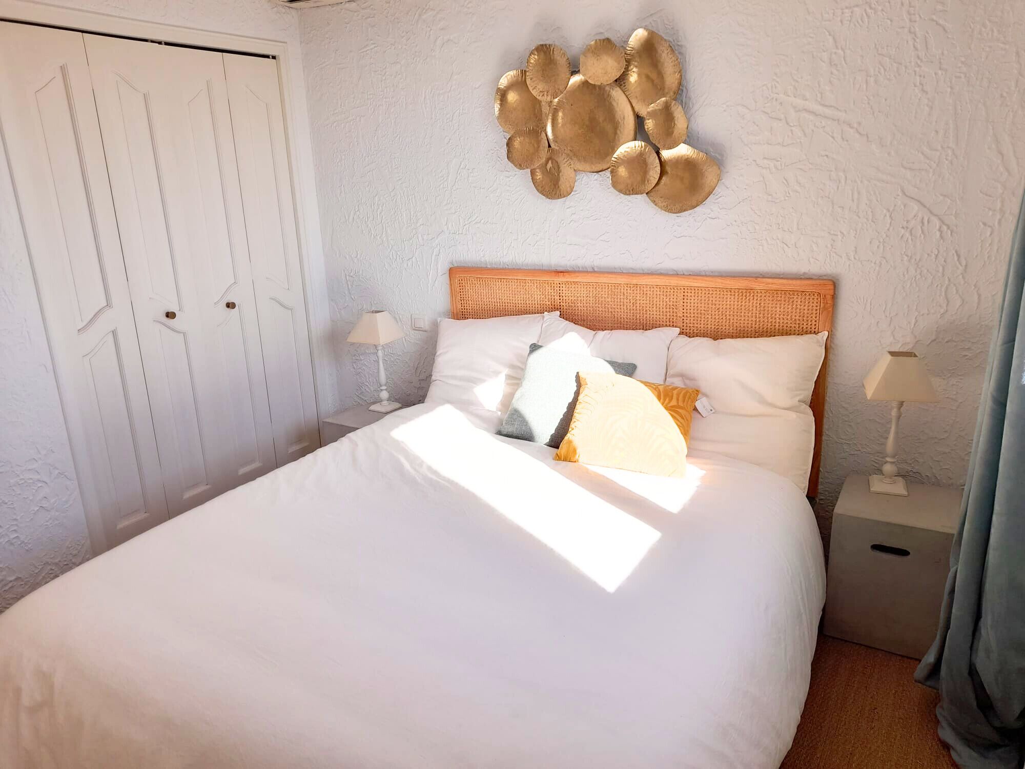 Un lit bien fait, avec une literie blanche, une tête de lit et plusieurs oreillers. On y voit deux tables de chevet avec des lampes, un élément décoratif fixé au mur et une porte au fond.