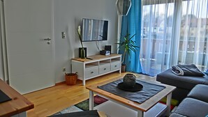Uma sala de estar moderna com uma televisão de tela plana, uma mesa de centro de madeira, um sofá cinza e uma janela com cortinas azuis.