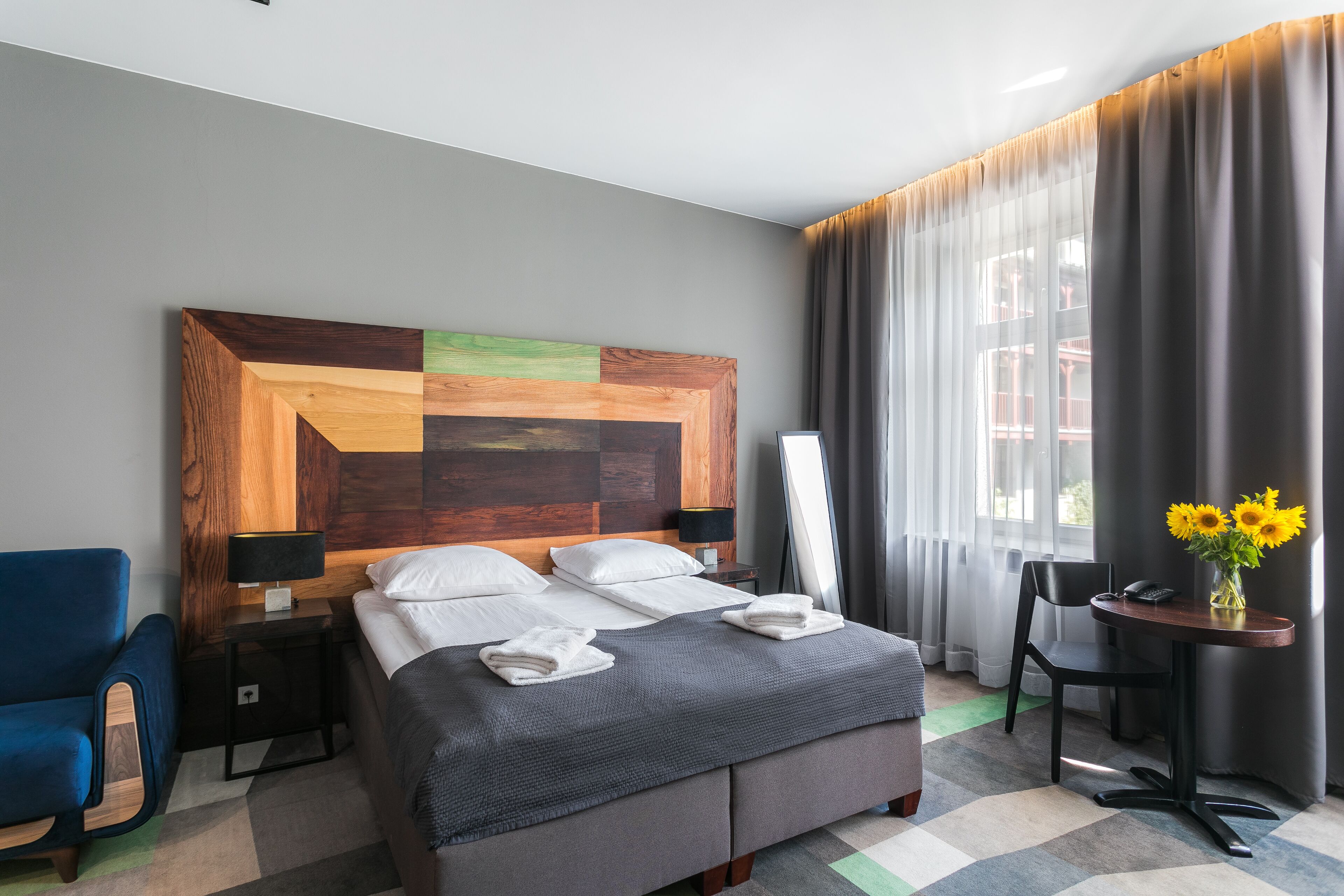 A hotel room with a large bed, a wooden headboard, a desk with a chair, a blue armchair, and a view of the outdoors through sheer curtains.