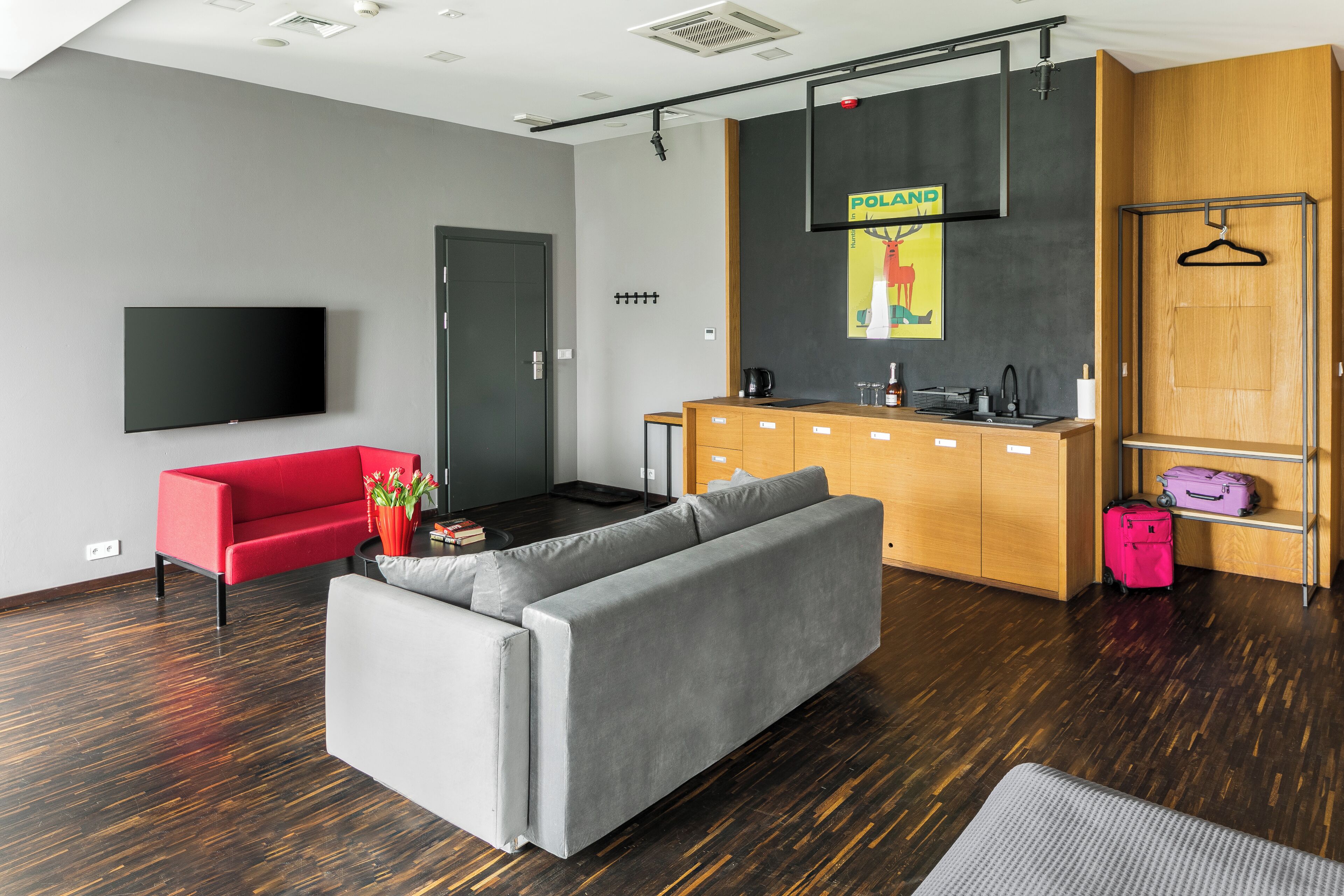 Ein modernes Wohnzimmer mit einem grauen Sofa, einem roten Sessel, einem Flachbildfernseher, einer Holzkommode und einer Küchenzeile mit einer Kaffeemaschine.