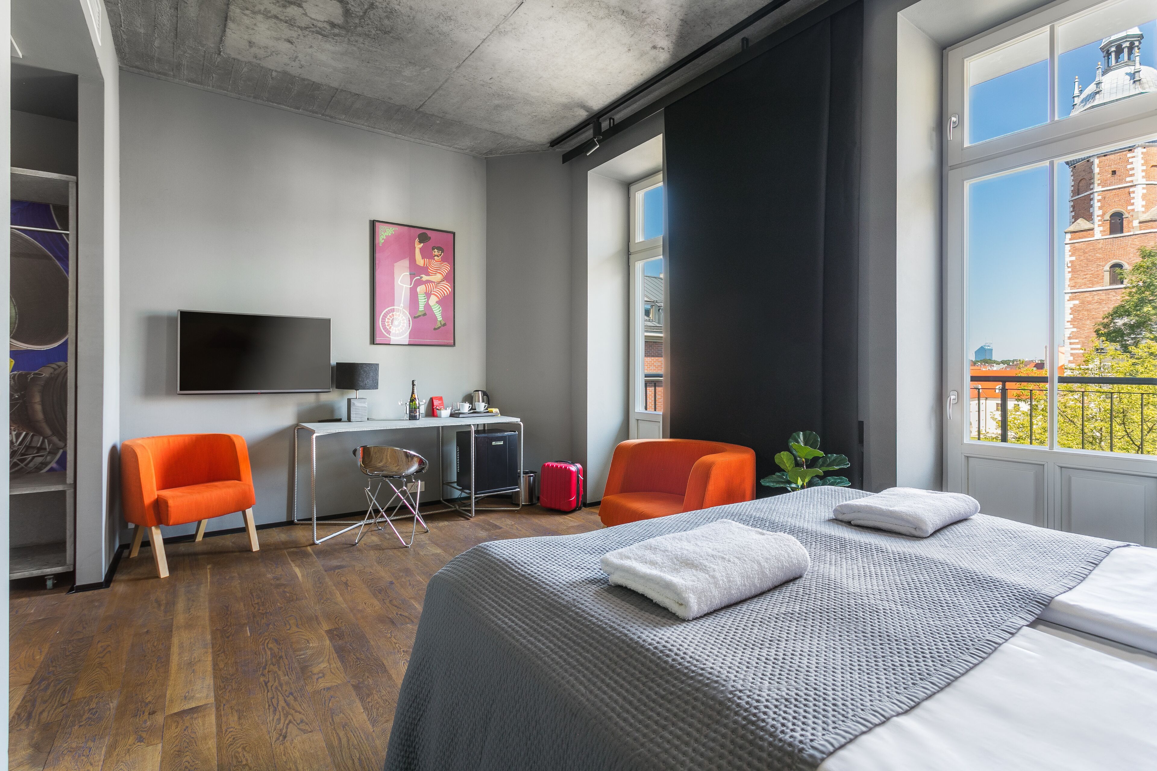 Ein Hotelzimmer mit einem Bett, zwei Sesseln, einem Schreibtisch, einem Fernseher und einem Fenster mit Blick auf Gebäude.