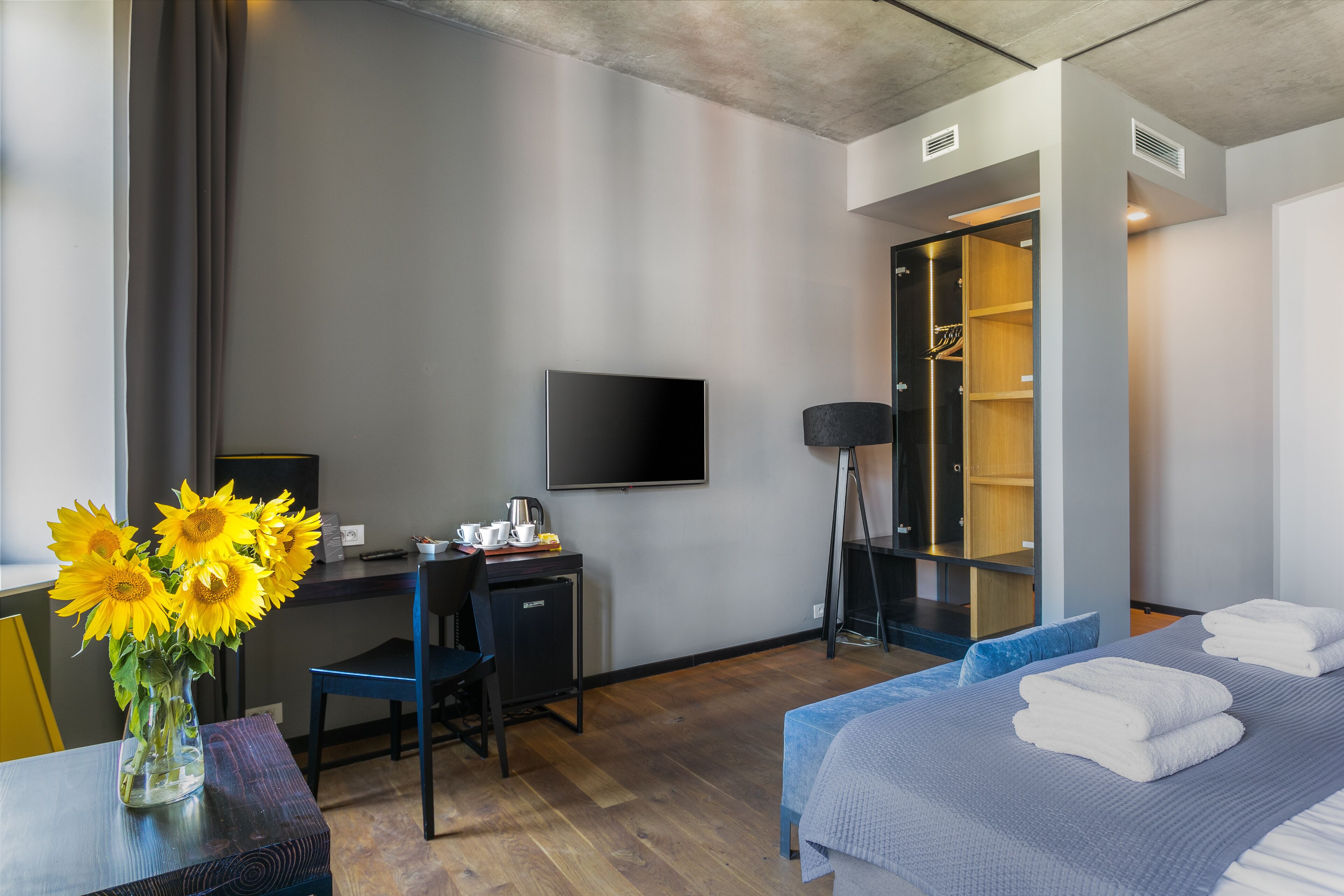 A hotel room with a bed, a desk with a chair, a television, a wardrobe, and a vase with sunflowers.