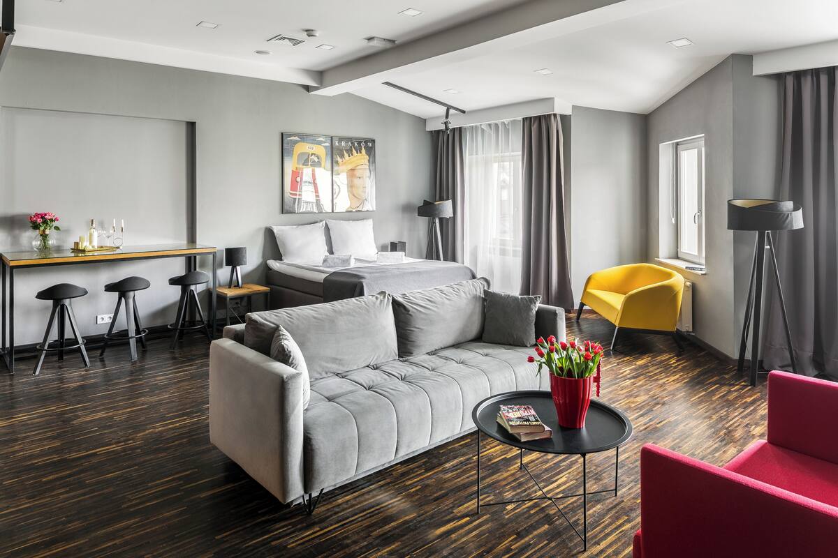 A modern hotel room with a grey sofa, a red armchair, a dining area with a table and stools, and a large window with curtains.