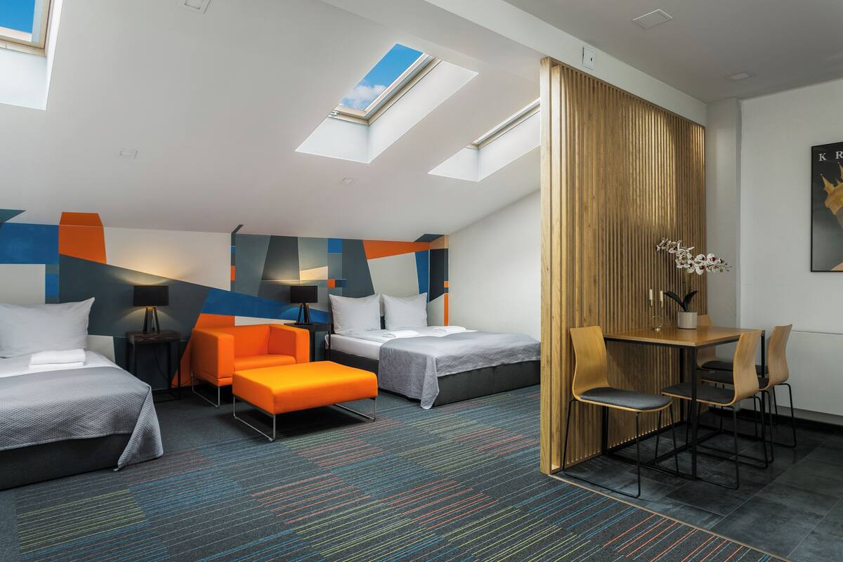 A hotel room with two beds, a desk, and a chair. There are skylights on the ceiling, a wooden partition, and a painting on the wall.