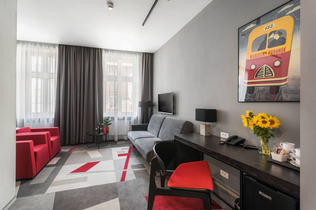 A modern hotel room with a grey sofa, red chairs, a TV, a desk with a vase of sunflowers, and a large poster on the wall.