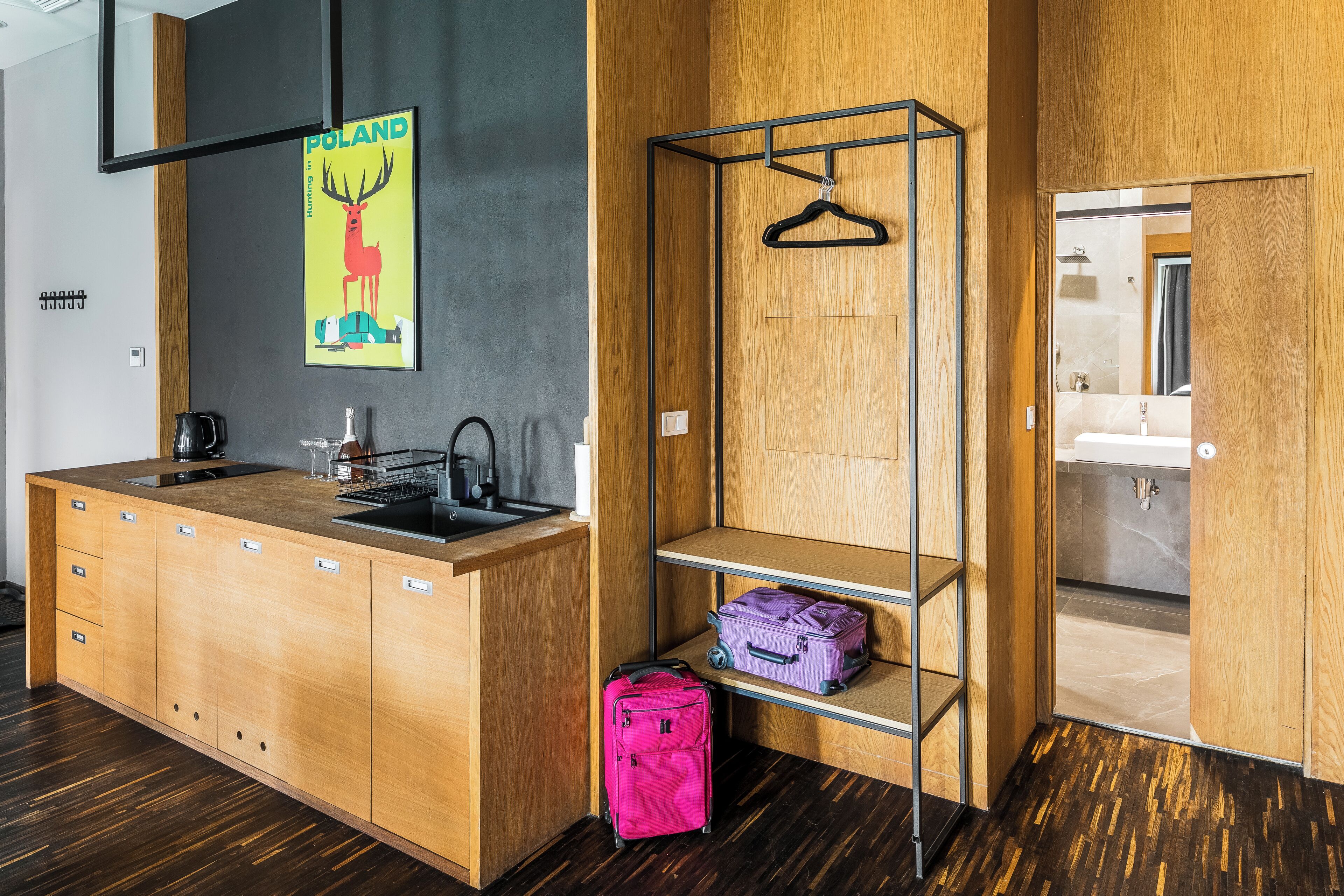 Une cuisine moderne avec un plan de travail en bois, un évier et une affiche représentant un cerf. Il y a une valise rose sur le sol et une valise violette sur une étagère.