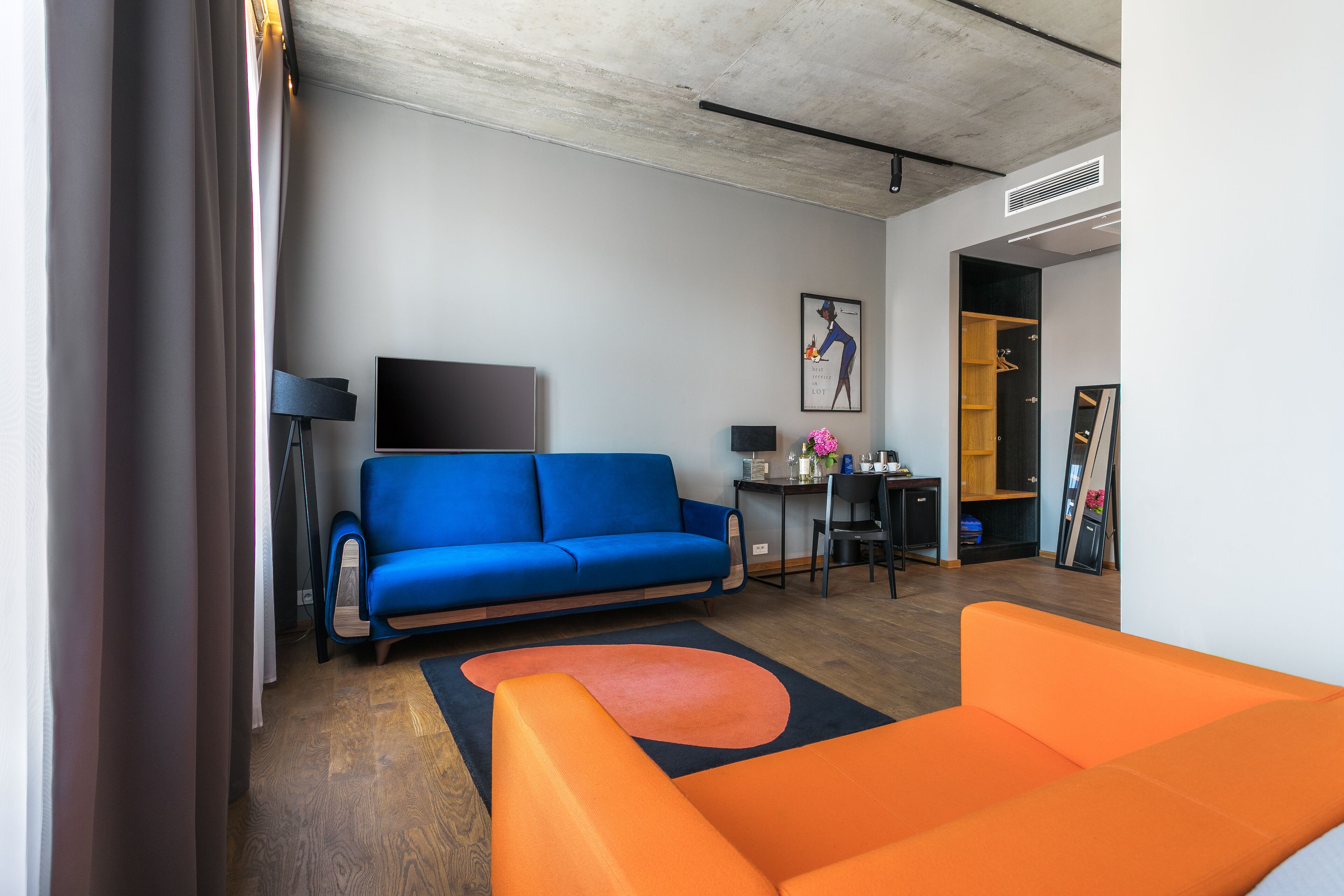 A modern living room with a blue sofa, an orange sofa, a flat-screen TV, a desk with a computer, and a framed picture on the wall.