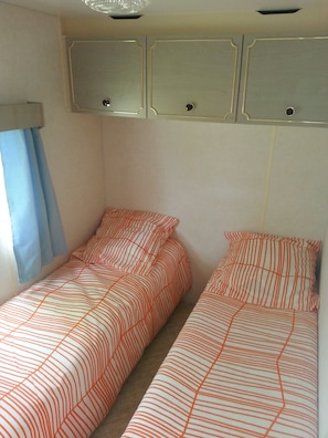 A small, compact bedroom with two single beds, each with striped bedding. There are overhead cabinets with two knobs, and a window with a blue curtain.