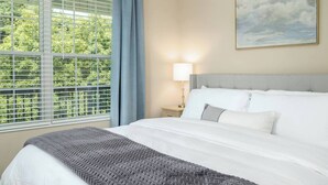 A hotel room with a large bed, a nightstand with a lamp, a window with blinds, and a painting on the wall.