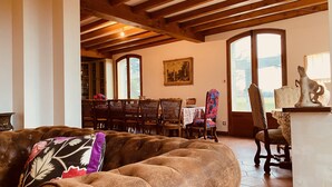 A cozy living room with a brown sofa, wooden ceiling beams, a dining area with wooden chairs, and a large window with a view.