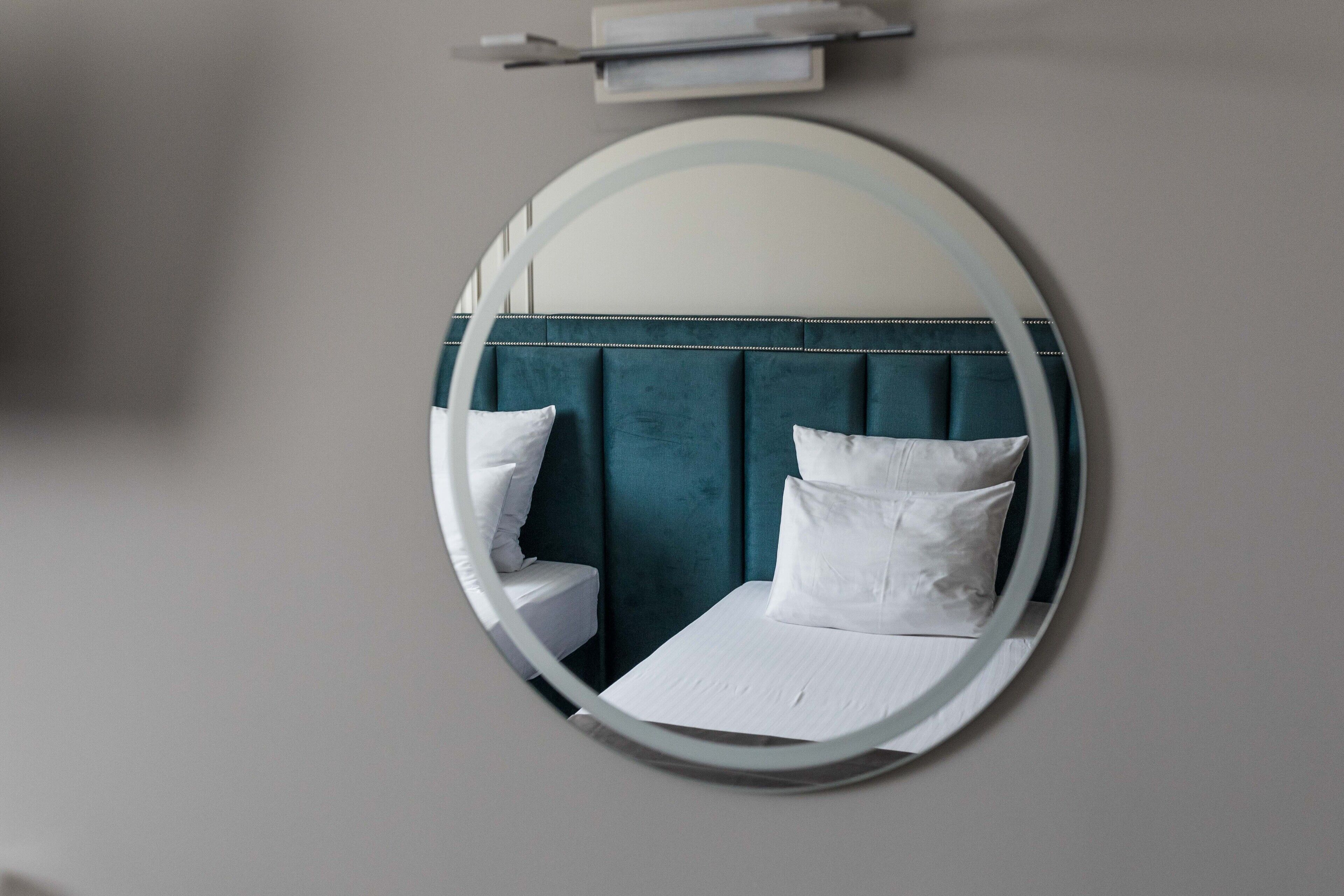 Un espejo circular refleja una cama con ropa blanca y dos almohadas, un cabecero tapizado en color turquesa y una mesita de noche blanca.