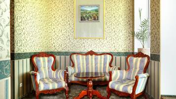 A room with patterned wallpaper, a framed landscape painting, two upholstered chairs, a wooden table, and a potted plant.