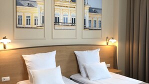 A hotel room with two beds, a wooden headboard, and wall-mounted artwork depicting historical buildings. - Anna Palace Hotel (Ruse)