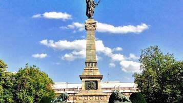 Ett högt monument med en staty på toppen, omgivet av ett stenlagt område med träd och en byggnad i bakgrunden.