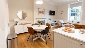 Ett modernt kök med ett matbord, stolar, en vit bänkskiva, en diskho, en spegel, en öppen spis, en tv och ett fönster med utsikt över grönområden.
