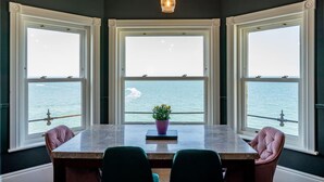A dining area with a table and chairs, large windows with a view of the sea, and hanging pendant lights. - Luxury Beachfront Penthouse, Panoramic Sea Views (Herne Bay)