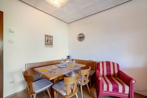 A dining area with a table set for four, wooden chairs, a red armchair, a wall clock, and a small decorative piece.
