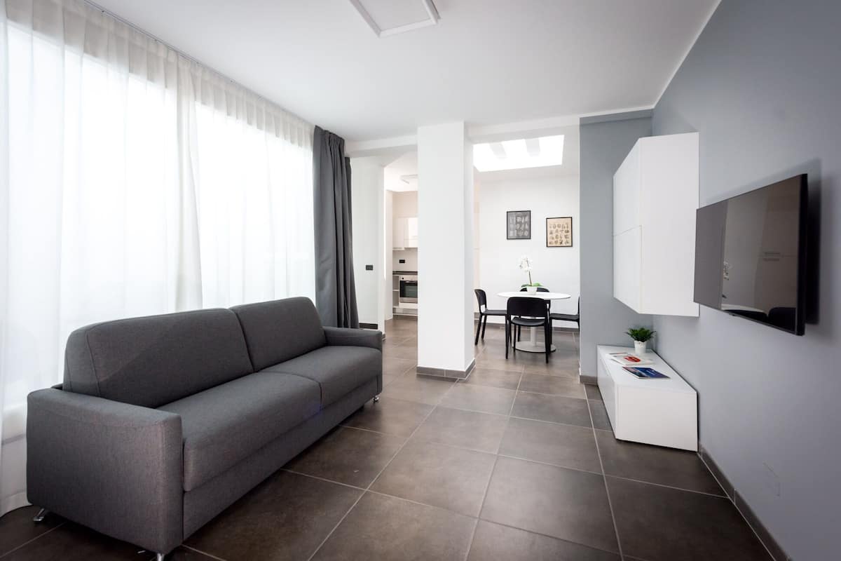 Una sala moderna con un sofá gris, un comedor con mesa y sillas, un televisor montado en la pared y una cocina al fondo.