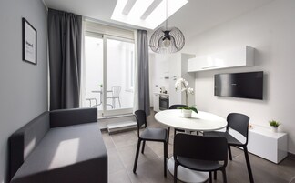 Una sala de estar moderna y bien iluminada con sofá, mesa de comedor y sillas, una zona de cocina y un amplio ventanal con cortinas.