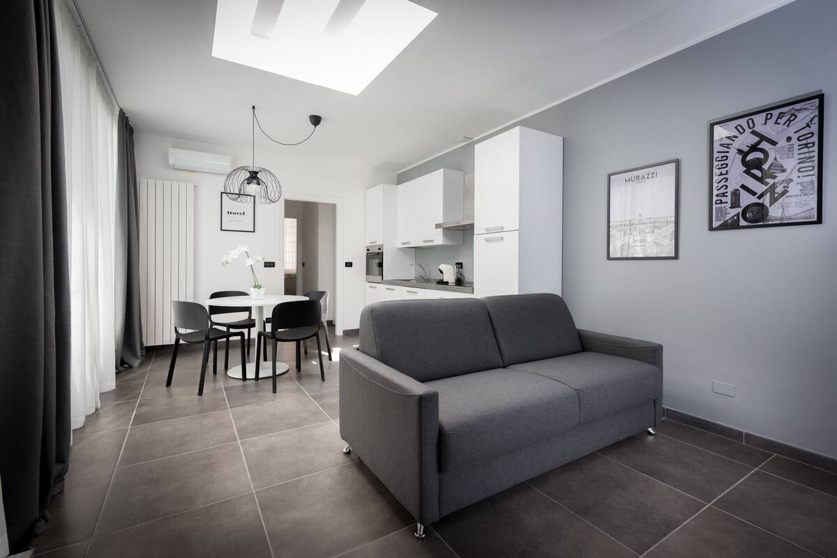 Una sala de estar moderna con un sofá gris, una mesa de comedor con sillas y una zona de cocina.