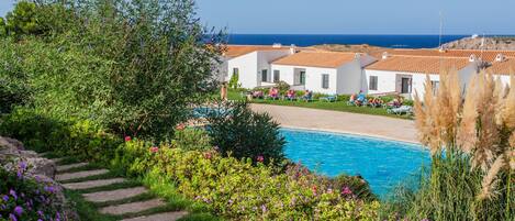 A poolside view of a resort with a clear blue sky, a swimming pool, and white buildings with red roofs.