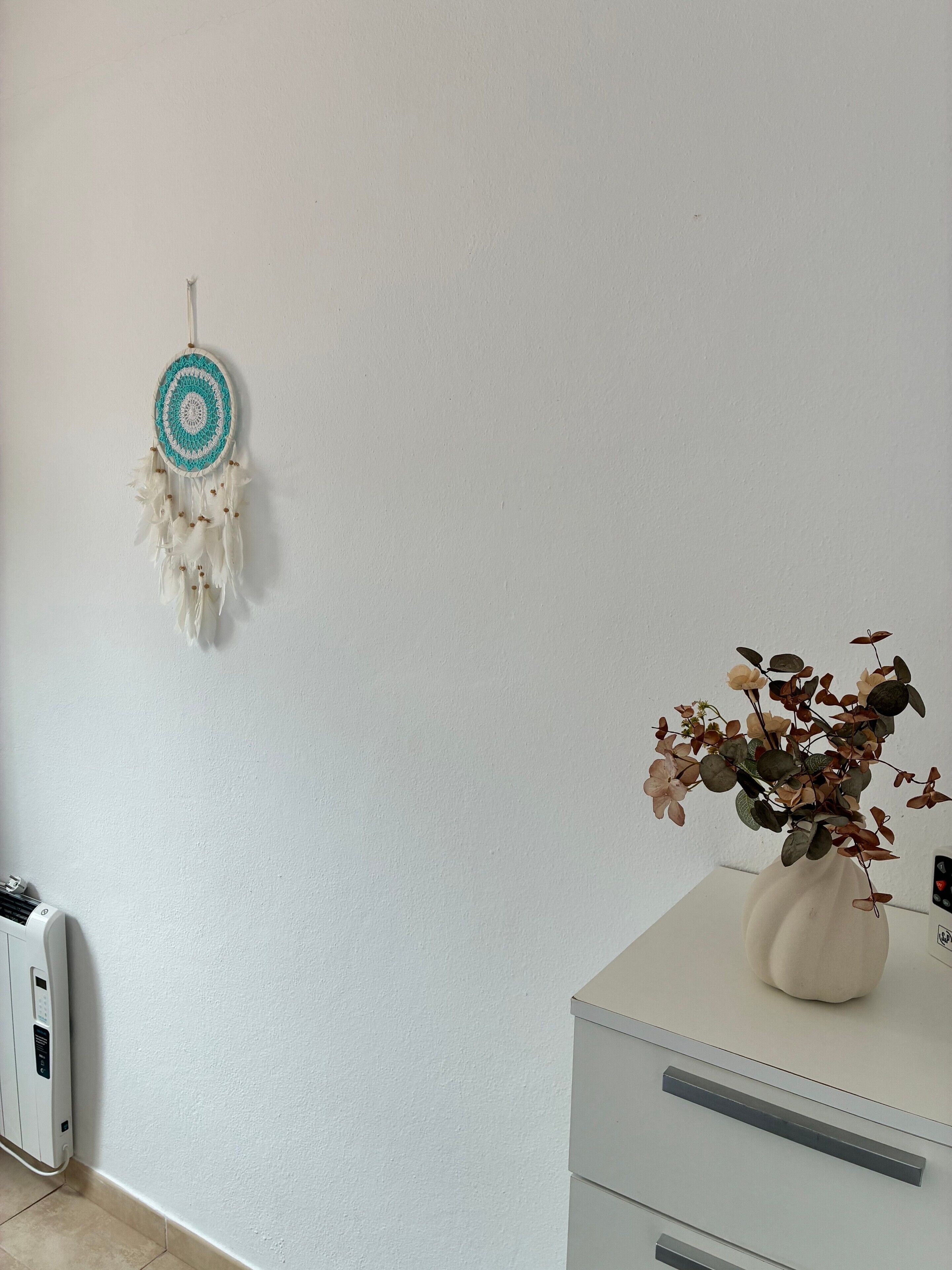 A room with a white wall, a white dresser with drawers, a white radiator, and a dreamcatcher with white and blue colors.