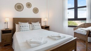 Ein ordentlich gemachtes Bett mit weißer Bettwäsche, zwei Nachttische mit Lampen, ein Korbstuhl und ein großes Fenster mit Vorhängen.