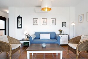 Una sala de estar con un sofá azul a cuadros, dos sillones de mimbre, una mesa de centro de madera y un equipo de aire acondicionado blanco en la pared.