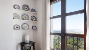 Una stanza con un'ampia finestra, uno scaffale a parete con piatti decorativi, un tavolino con una pianta in vaso e una porta che si affaccia su un paesaggio.