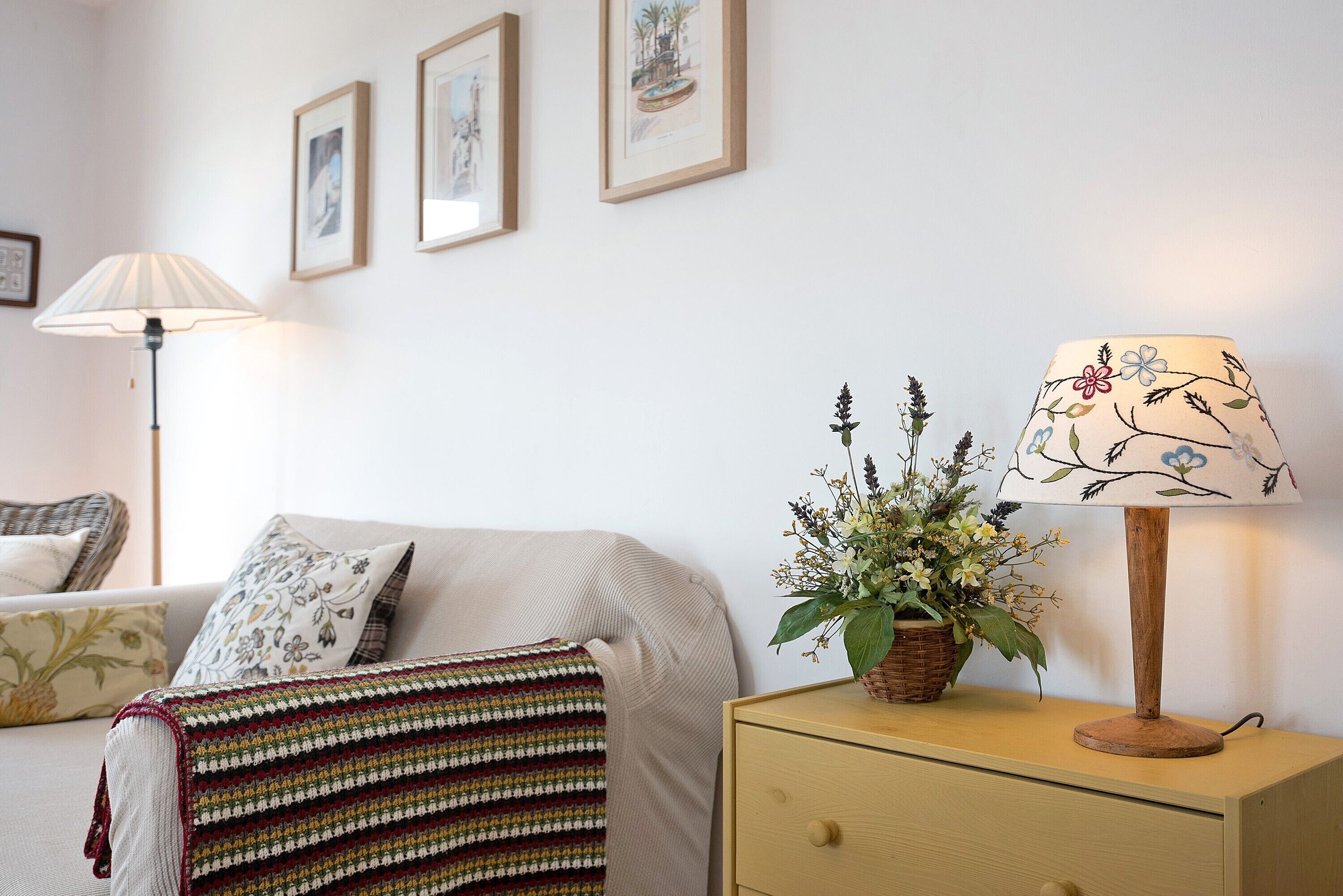 A bedroom with a bed, a nightstand with a lamp, a wall-mounted shelf, and framed artwork.