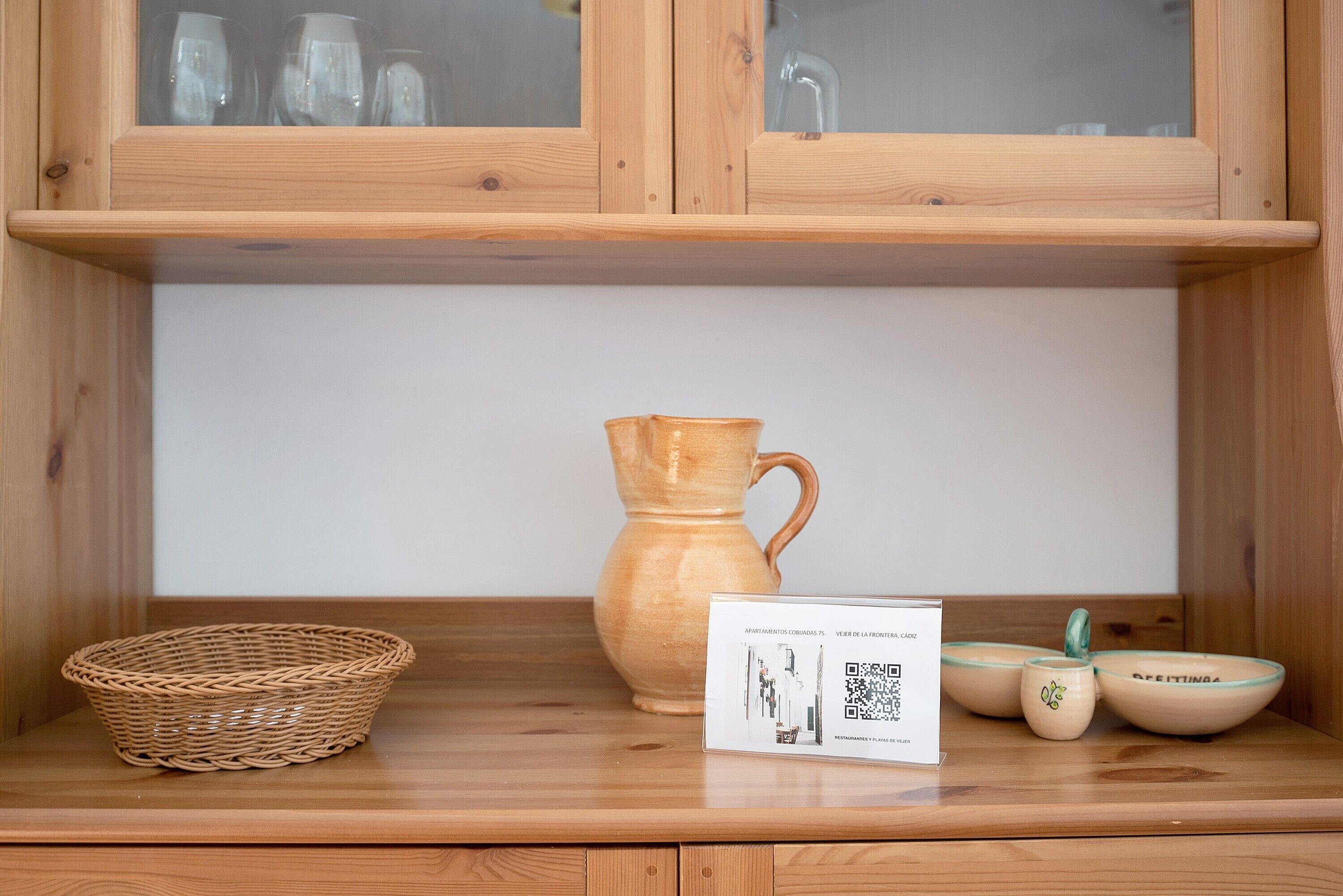 Un armadietto in legno con ante in vetro, un cesto intrecciato, un boccale in ceramica e ciotole in ceramica su una mensola in legno.
