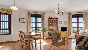 A dining area with a table and chairs, a wicker sofa, a television, and a cabinet with kitchenware. - Apartment Cobijadas75 Segur with Mountain View, Balcony & Wi-Fi (Vejer de la Frontera)