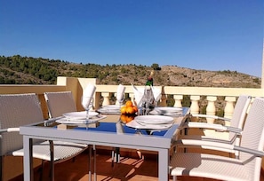 A dining table set for four with white chairs, a glass table, and a view of a hilly landscape.