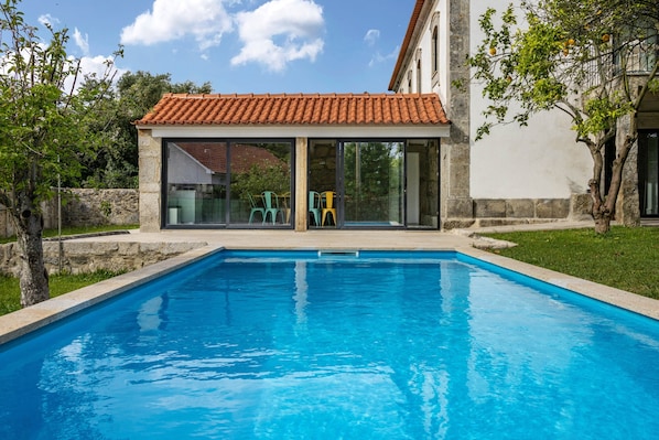 Une piscine dotée d’une structure de toit en carrelage, de murs en pierre et d’une zone herbeuse avec des arbres.
