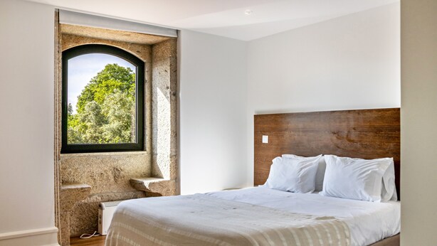 A bedroom with a large bed, a window with a view of trees, a stone wall, and a wooden headboard. - Holiday Apartment 'Casa do Mestre T2' with Mountain View, Pool and Wi-Fi (Vila de Punhe)