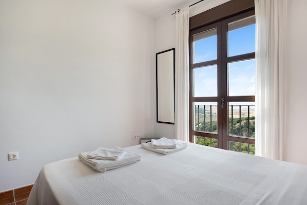 A bedroom with a large window, a bed with white linens, and a view of greenery outside.