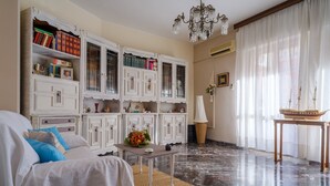 Une pièce avec un canapé blanc, une petite table sur laquelle est posé un vase, une bibliothèque, un lustre et une maquette de bateau sur une table.