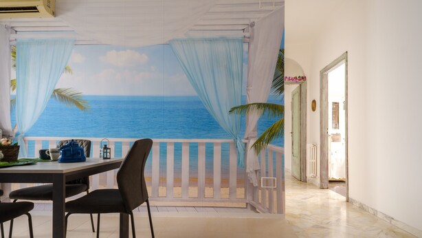 Un espace repas offrant une vue sur l’océan grâce à une fresque murale, une balustrade blanche et une porte donnant sur une autre pièce.