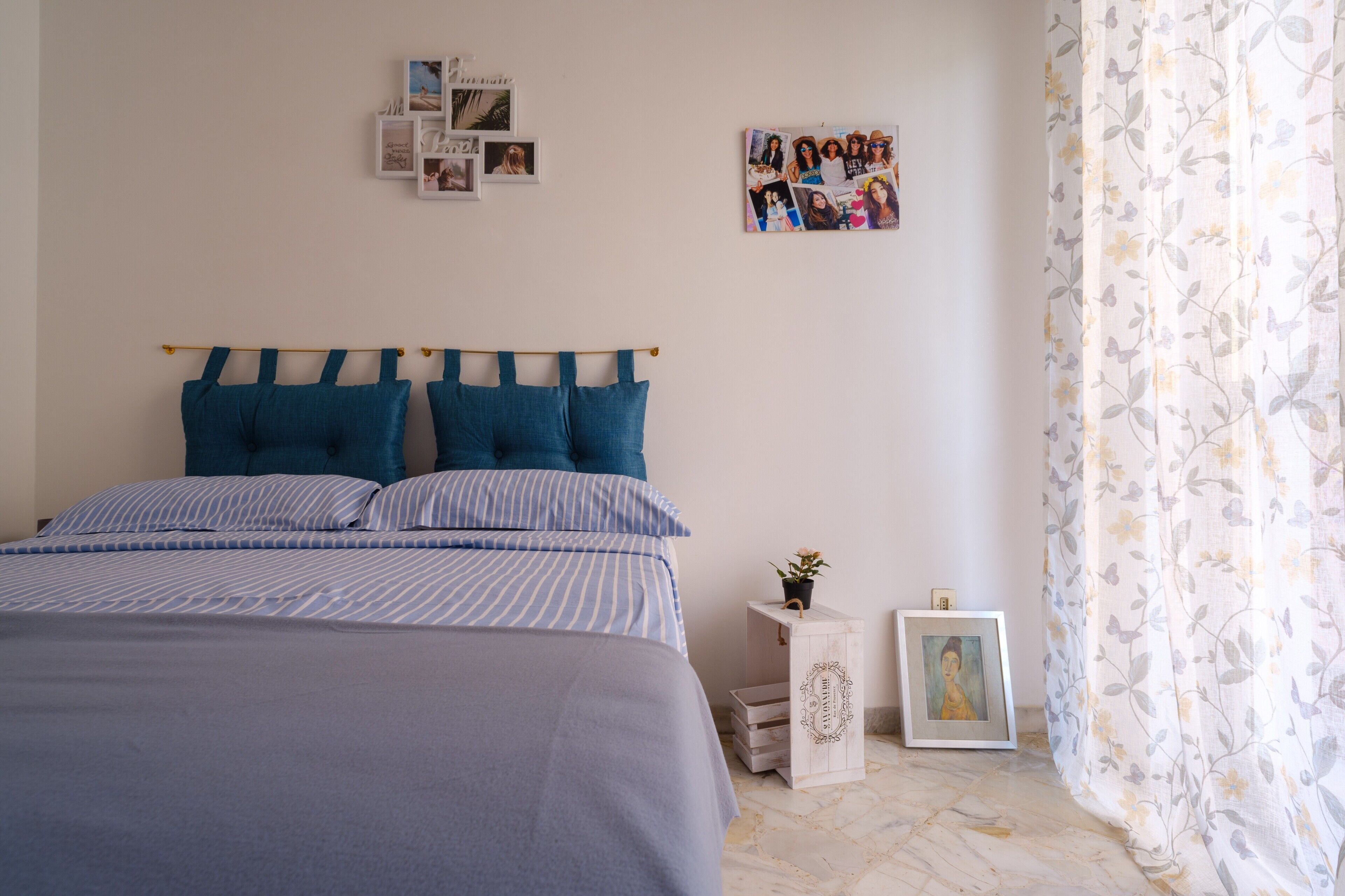 A bedroom with a bed, pillows, a bedside table with a plant, a framed picture, and a wall with photos.