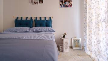A bedroom with a bed, pillows, a bedside table with a plant, a framed picture, and a wall with photos.