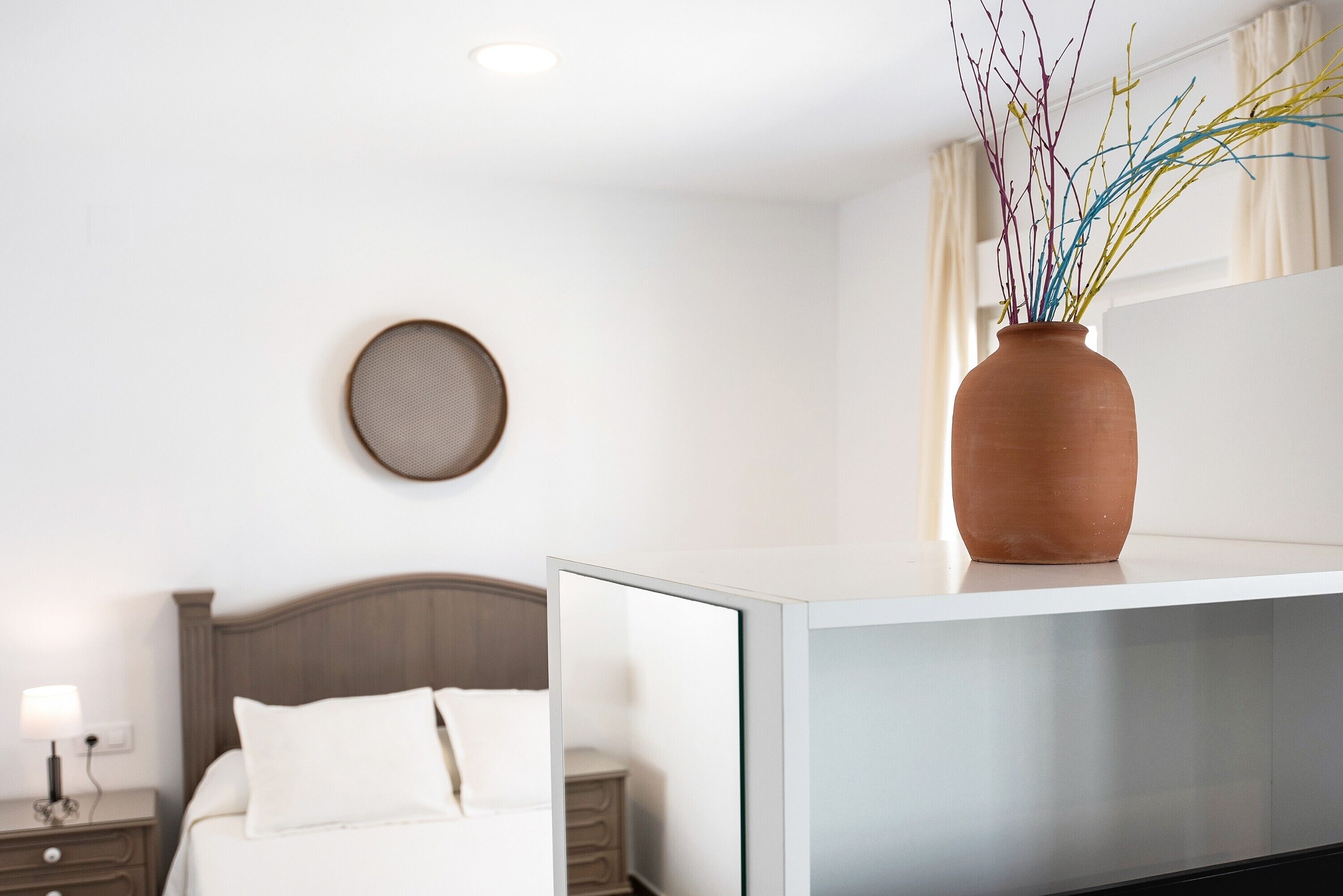 A hotel room with a bed, bedside table, lamp, and a vase with branches on a dresser.
