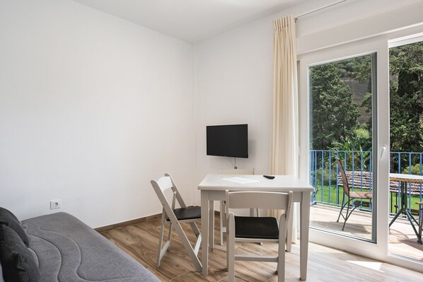 A room with a sofa, a table, chairs, a TV, and a balcony with a view of greenery.