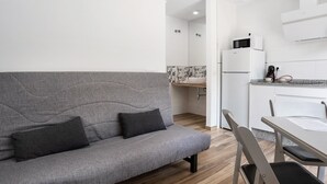 A grey sofa with two cushions, a wooden floor, a white kitchen with a microwave and a refrigerator, and a small table with chairs.