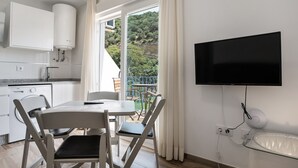 A modern kitchen with a built-in dishwasher, a dining area with a table and chairs, a flat-screen TV mounted on the wall, and a balcony with a view of greenery.