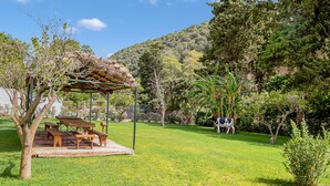 A covered outdoor seating area with benches and a table, surrounded by lush greenery and a hill in the background.