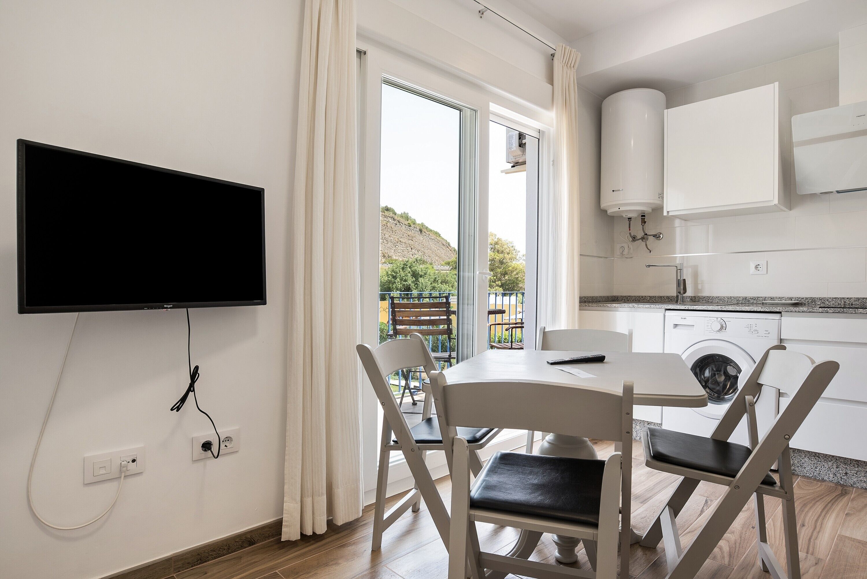 Une cuisine moderne équipée d’un lave-linge intégré, un coin repas avec une table et des chaises, et une grande fenêtre avec des rideaux.