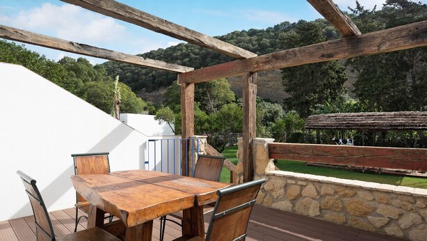 An outdoor wooden deck with a table and chairs, stone wall, and a view of a green landscape.