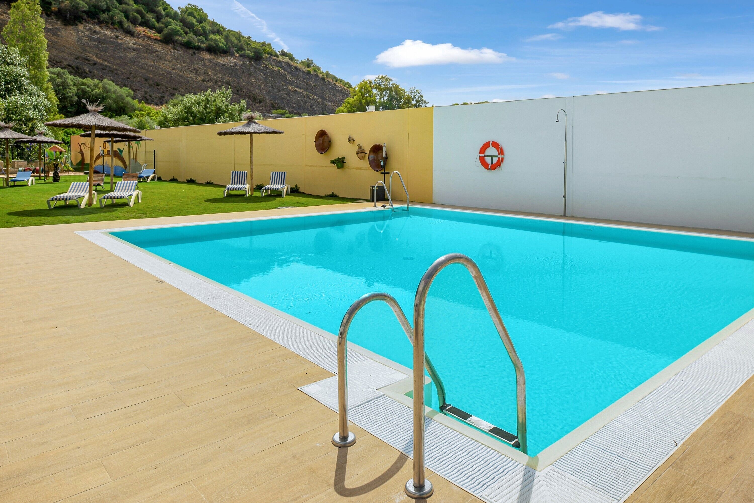 Une piscine avec une terrasse en bois, des chaises longues et un panneau indiquant la présence d’un sauveteur.