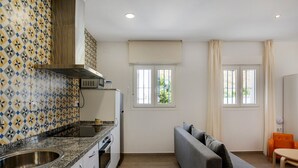 Una cocina moderna con horno empotrado, un fregadero y un microondas. Hay dos ventanas con cortinas, un sofá y una pequeña mesa.