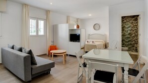 Una habitación moderna y bien iluminada con sofá, mesa de comedor, sillas, televisor, cama y baño con pared de mosaicos.
